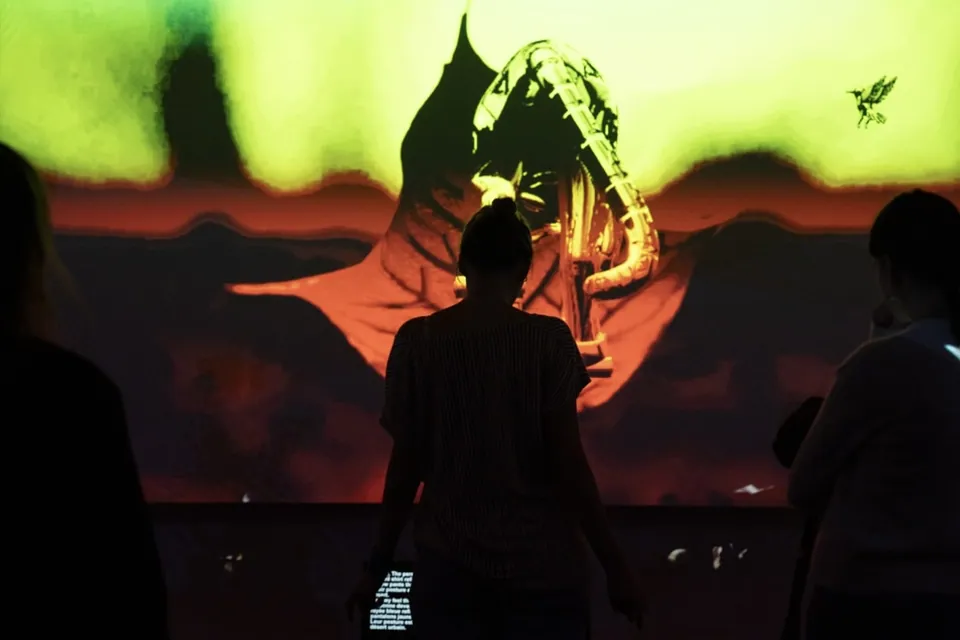 Visitors in silhouette before a projection of a jaguar figure against a red and green sky.