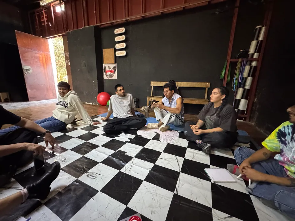 Dancers and the lab team sit along a wall and on the checkered floor, listening.