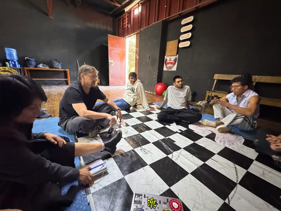 The team talks in a circle on the checkered floor, around a laptop and sheets of paper.