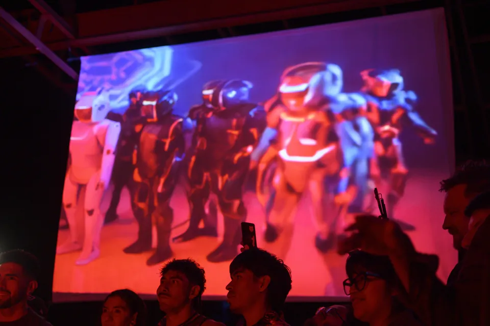 A screen shows generated figures in glowing armour, red and blue, above the audience.
