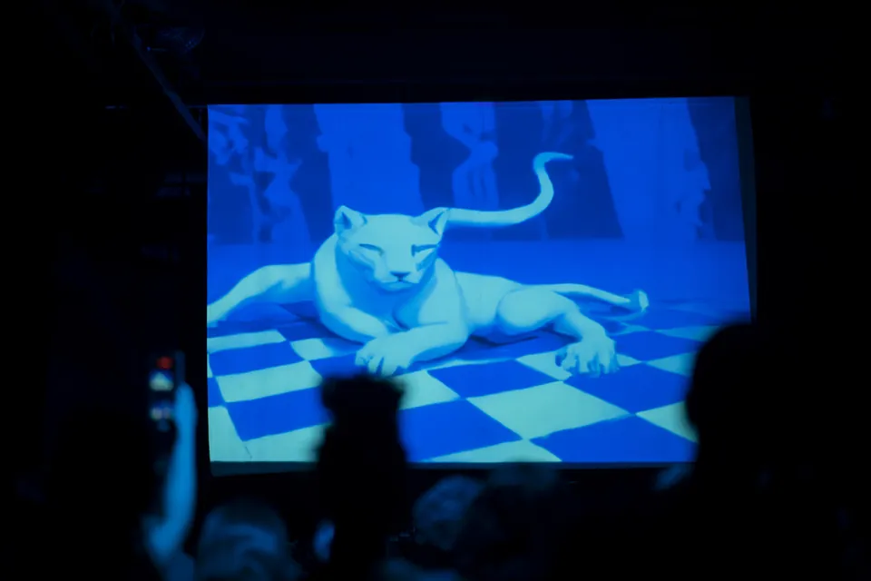 A screen shows a generated white jaguar lying on a checkered floor, above the silhouettes of the audience.