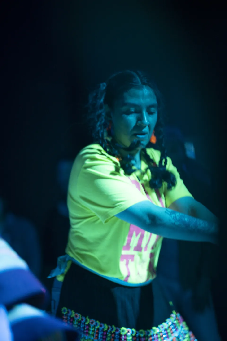 A dancer in a bright yellow shirt and a beaded skirt moves under blue light.