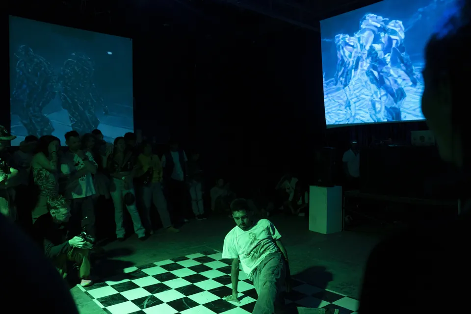 A dancer on a lit checkered floor in a dark hall, the audience in a ring, screens above.