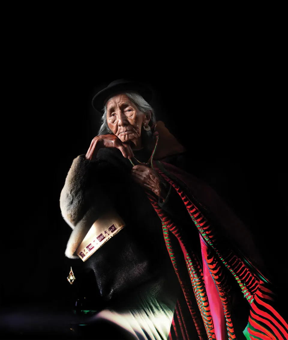 A portrait from Las Awichas: an elderly woman in a dark hat rests her chin on her hand. Her shawl turns into red and green stripes.