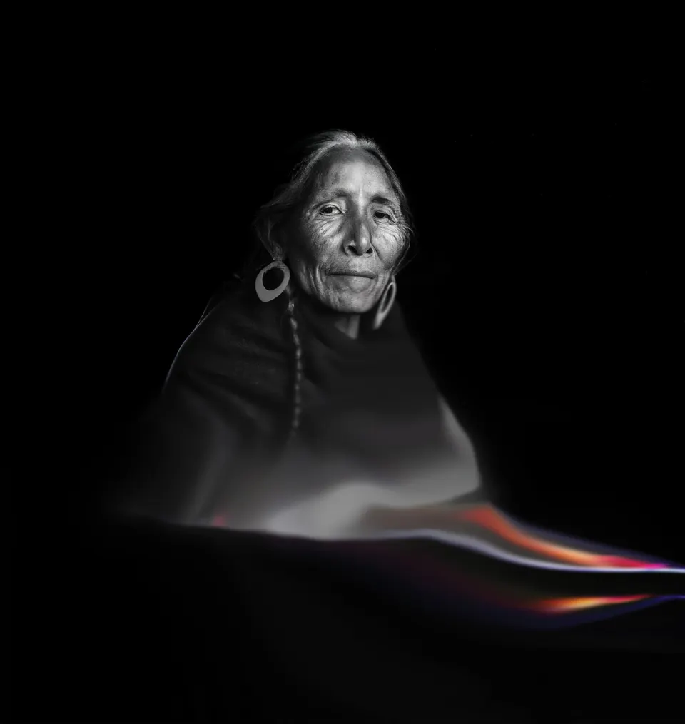 A portrait from Las Awichas in black and white: an elderly woman with a braid and a large earring. A band of colour runs below her.