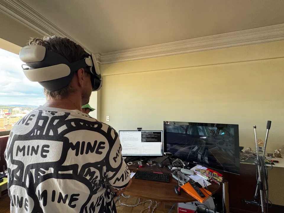A person wearing a VR headset stands in a room with a desk, monitors and a window over the city. Seen from behind.
