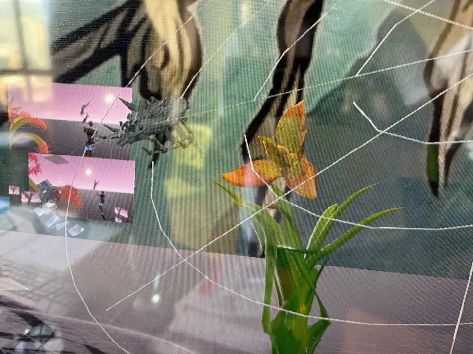 A 3D spider on its web beside an orange flower. Two small windows show a pink virtual world.